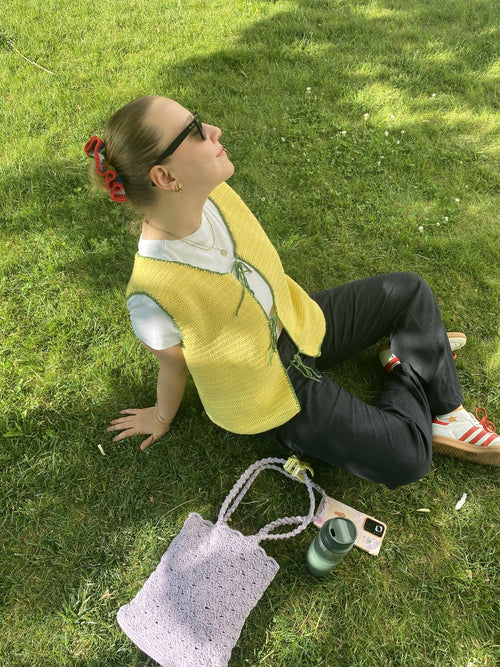 Customer sitting on grass in a yellow crochet vest with a crochet bag