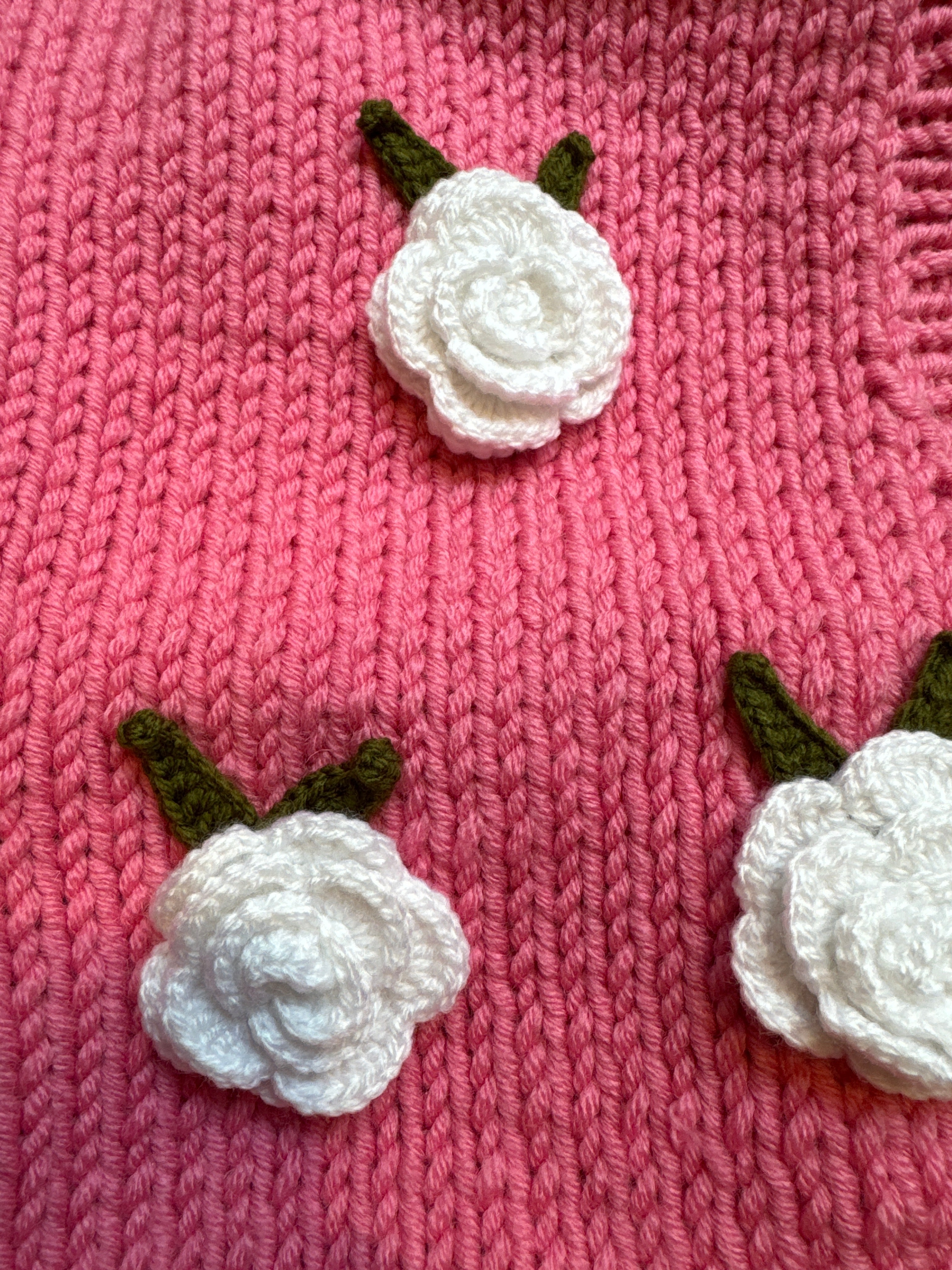 Close-up of crocheted white roses with green leaves on pink chunky knit — handmade by Ana Chic