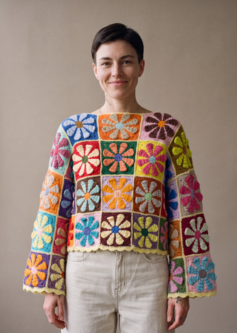 Woman wearing the Flower Jumper, a handmade crochet granny-square jumper with flat daisy motifs, boat neckline and wide bell sleeves, styled with ecru jeans