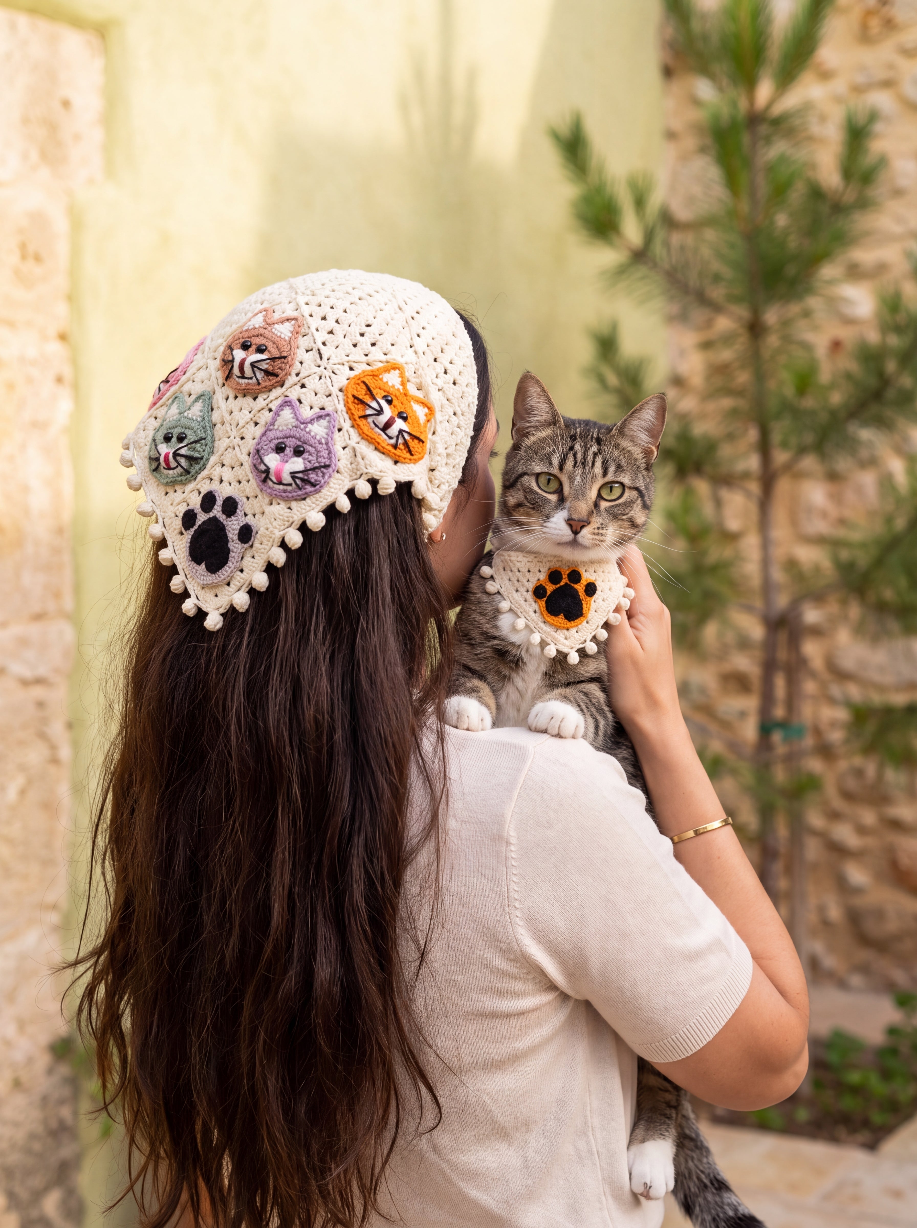 Woman wearing the large cat-face crochet bandana over her hair, holding a tabby cat in the matching small paw bandana — handmade by Ana Chic