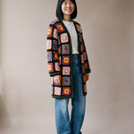 Full-length view of the long black granny square crochet cardigan ending just above the knee, worn with wide-leg jeans, size S