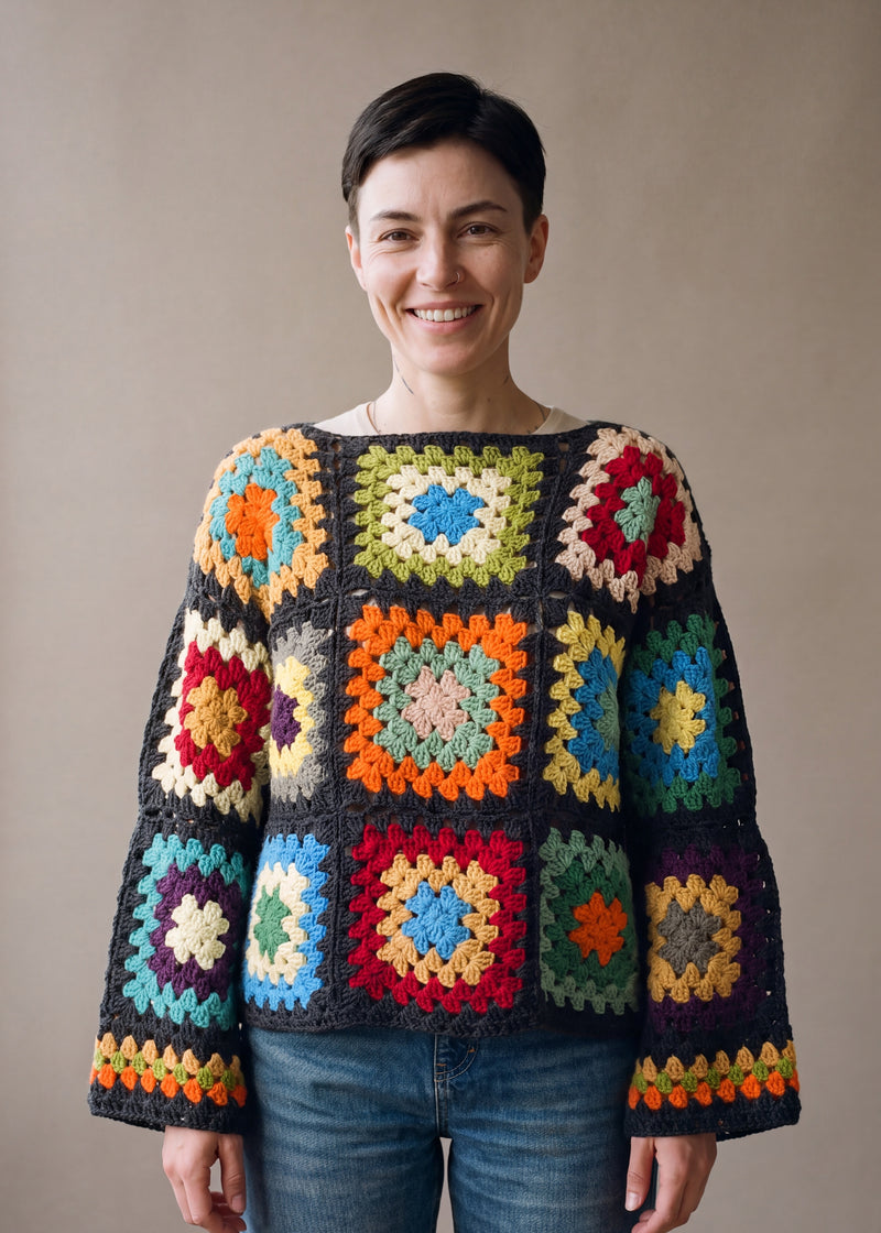 Woman wearing the Retroa jumper, a handmade crochet granny-square jumper in bright colours joined in charcoal, with boat neckline and wide sleeves, styled with blue jeans