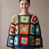 Woman wearing the Retroa jumper, a handmade crochet granny-square jumper in bright colours joined in charcoal, with boat neckline and wide sleeves, styled with blue jeans